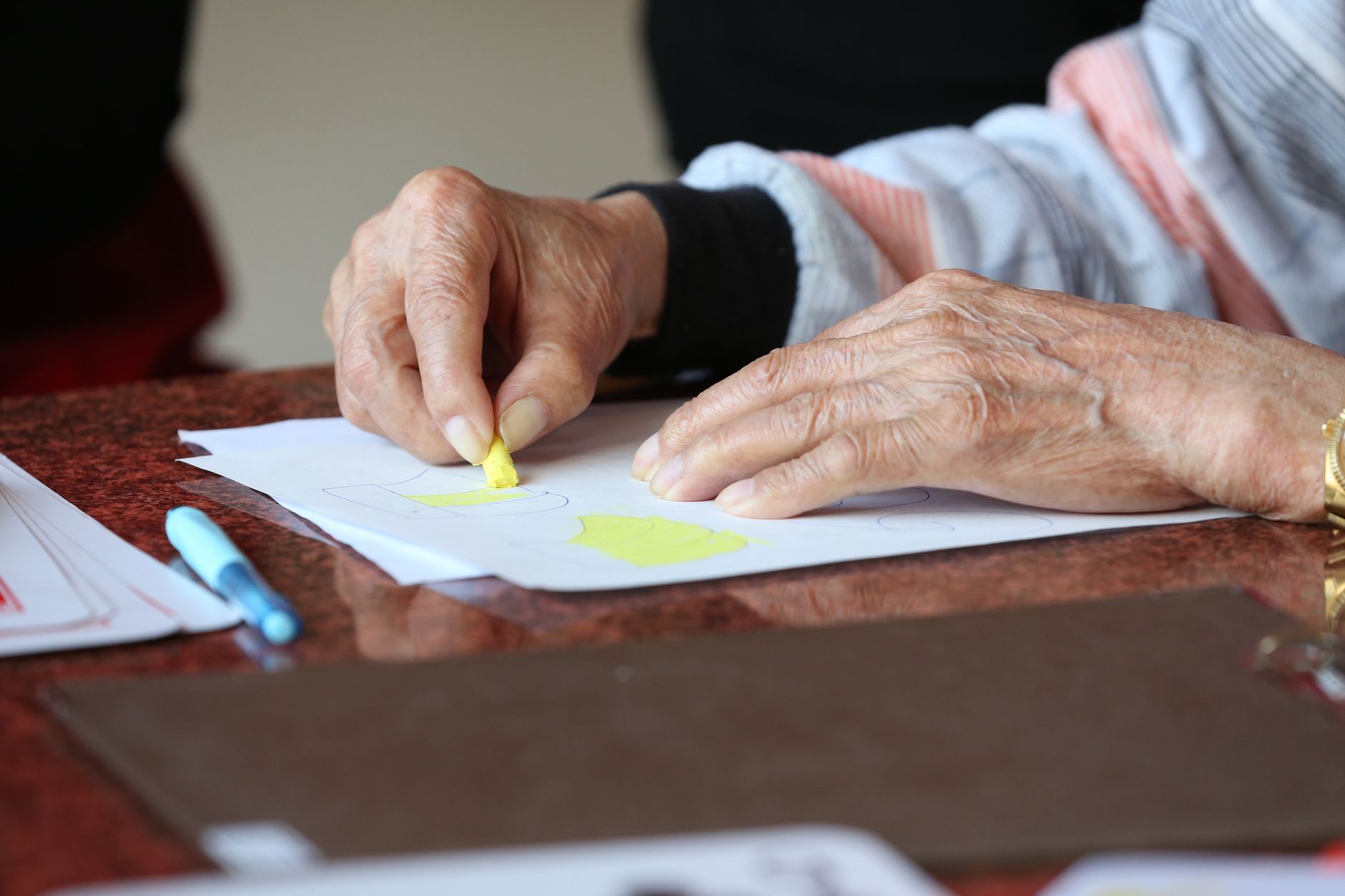 Ältere Person malt konzentriert mit einem gelben Wachsmalstift am Tisch
